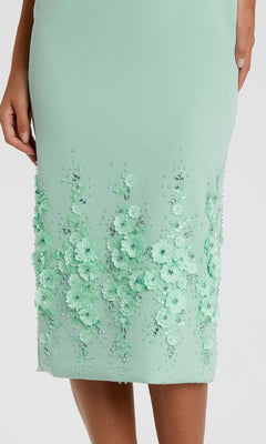 A close-up of the mint green Mac Duggal 11853 midi sheath wedding guest dress, featuring 3D floral appliqués and bead embellishments near the hem, worn by a standing model.