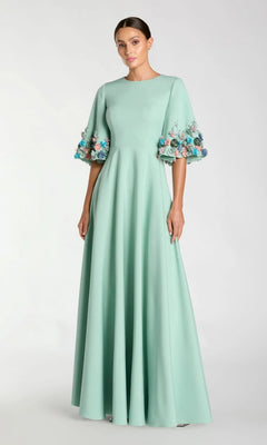 A woman in the Mac Duggal 11854 Bell-Sleeve Long Formal Dress—a pastel green, floor-length evening gown with multicolored floral embellishments—stands against a plain background.