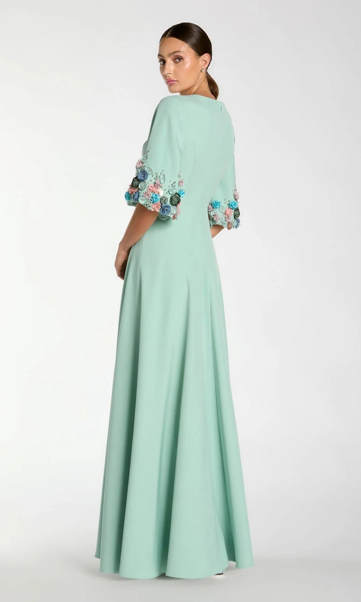 Woman wearing the Bell-Sleeve Long Formal Dress: Mac Duggal 11854, a floor-length, light green modest gown with floral embroidery on the bell sleeves, standing and looking over her shoulder.