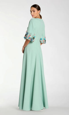 Woman wearing the Bell-Sleeve Long Formal Dress: Mac Duggal 11854, a floor-length, light green modest gown with floral embroidery on the bell sleeves, standing and looking over her shoulder.