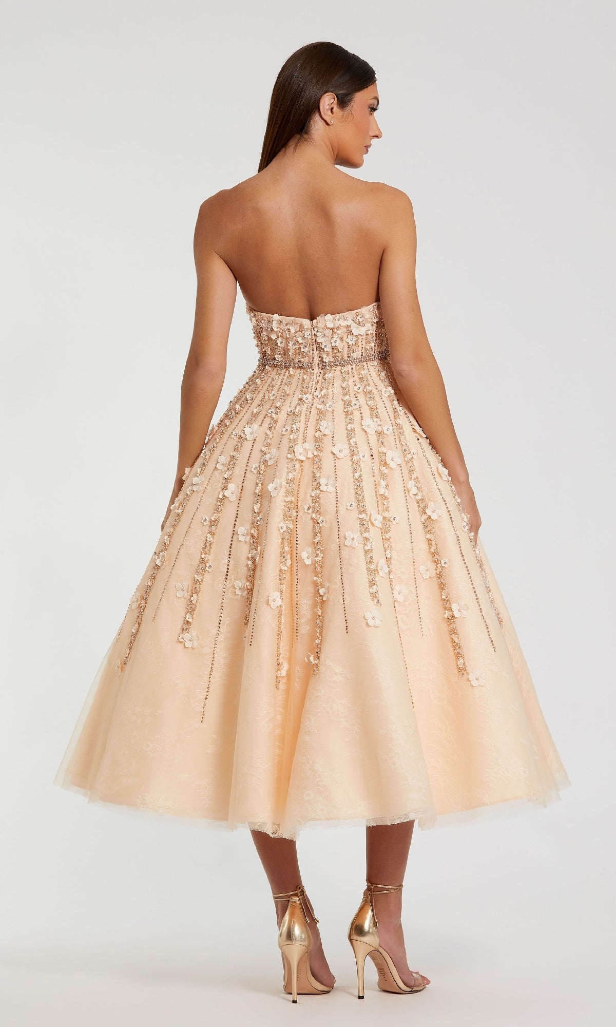 Wearing the Mac Duggal 11865 Strapless Champagne Party Dress, a woman in heels stands with her back to the camera; the tea-length, floral-embellished peach midi dress is ideal for semi-formal events.