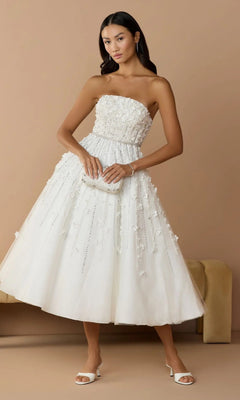A woman wears the Mac Duggal 11865 Strapless Ivory Party Dress, featuring floral embellishments, paired with a matching clutch and white heels, as she poses against a beige background.