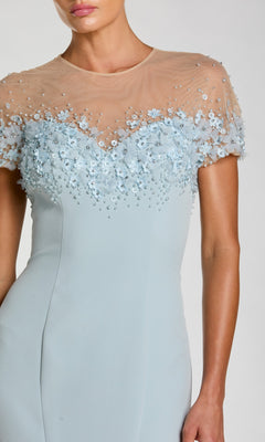 A woman wears the Mac Duggal 11867 short sleeve long formal dress in light blue, featuring a sheer nude mesh top embellished with small blue flowers and beads; only her upper body is visible.