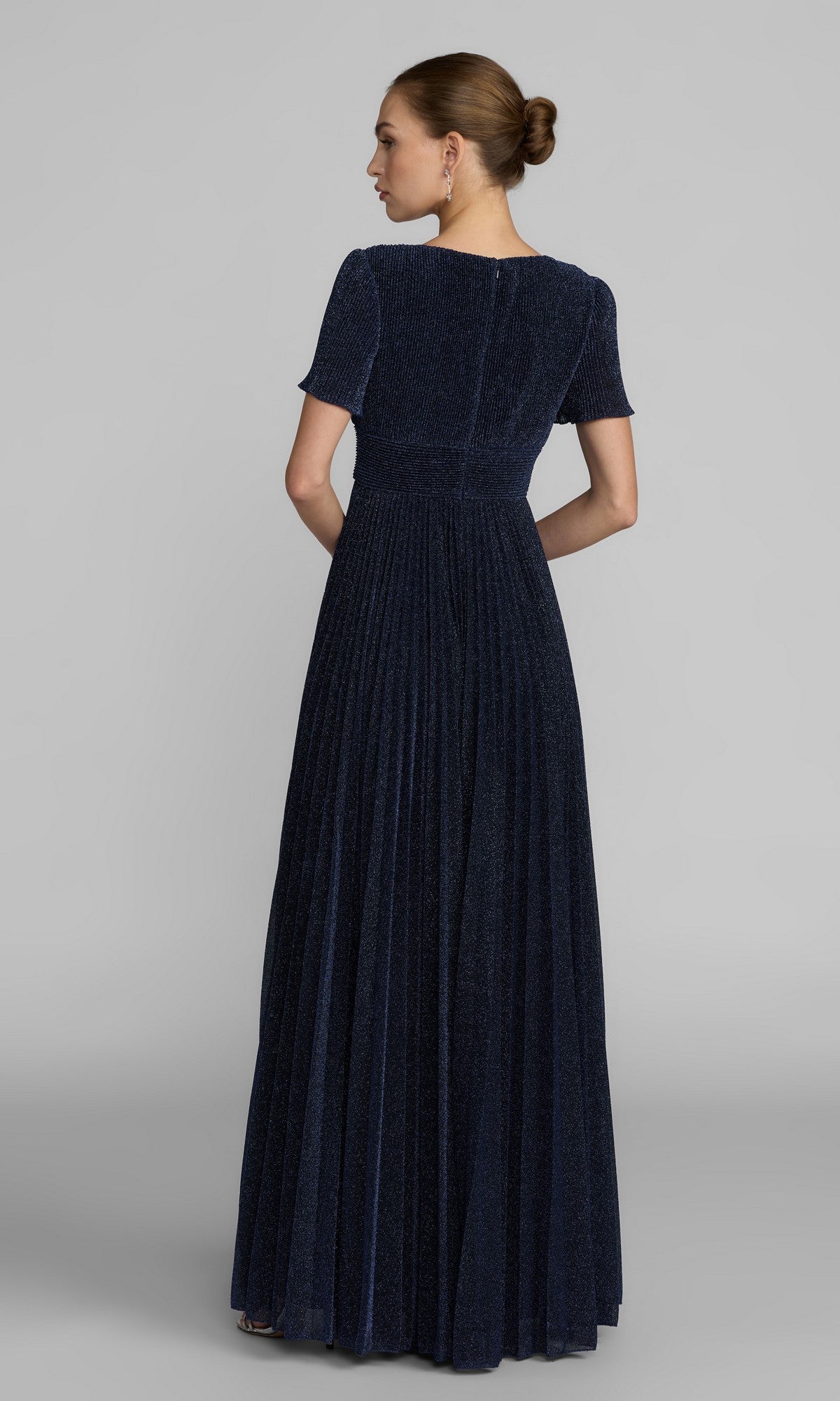 A person faces away, wearing the Mac Duggal 11881 Short Sleeve Pleated Formal Gown in dark blue with a floor-length skirt. Their hair is in a low bun, set against a plain gray background.