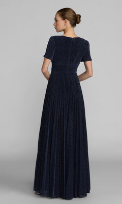 A person faces away, wearing the Mac Duggal 11881 Short Sleeve Pleated Formal Gown in dark blue with a floor-length skirt. Their hair is in a low bun, set against a plain gray background.