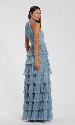 A woman wearing the Mac Duggal 11882 long formal dress—a sleeveless, light blue tiered gown with a halter neckline—stands facing away and looks over her shoulder against a plain white background.
