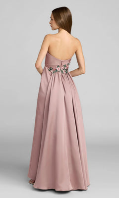 A woman stands facing away in the Mac Duggal 11894—a strapless, floor-length dusty pink gown with floral embroidery at the waist, capturing a timeless elegance characteristic of Mac Duggal’s signature formal style.