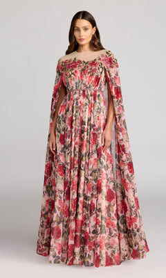 A woman models the Mac Duggal 11907 long formal dress in Rose Multi, featuring a floor-length chiffon design with elegant cape sleeves. The gown showcases a gathered bodice and delicate floral prints in shades of red, pink, and green, beautifully set against a light gray background.