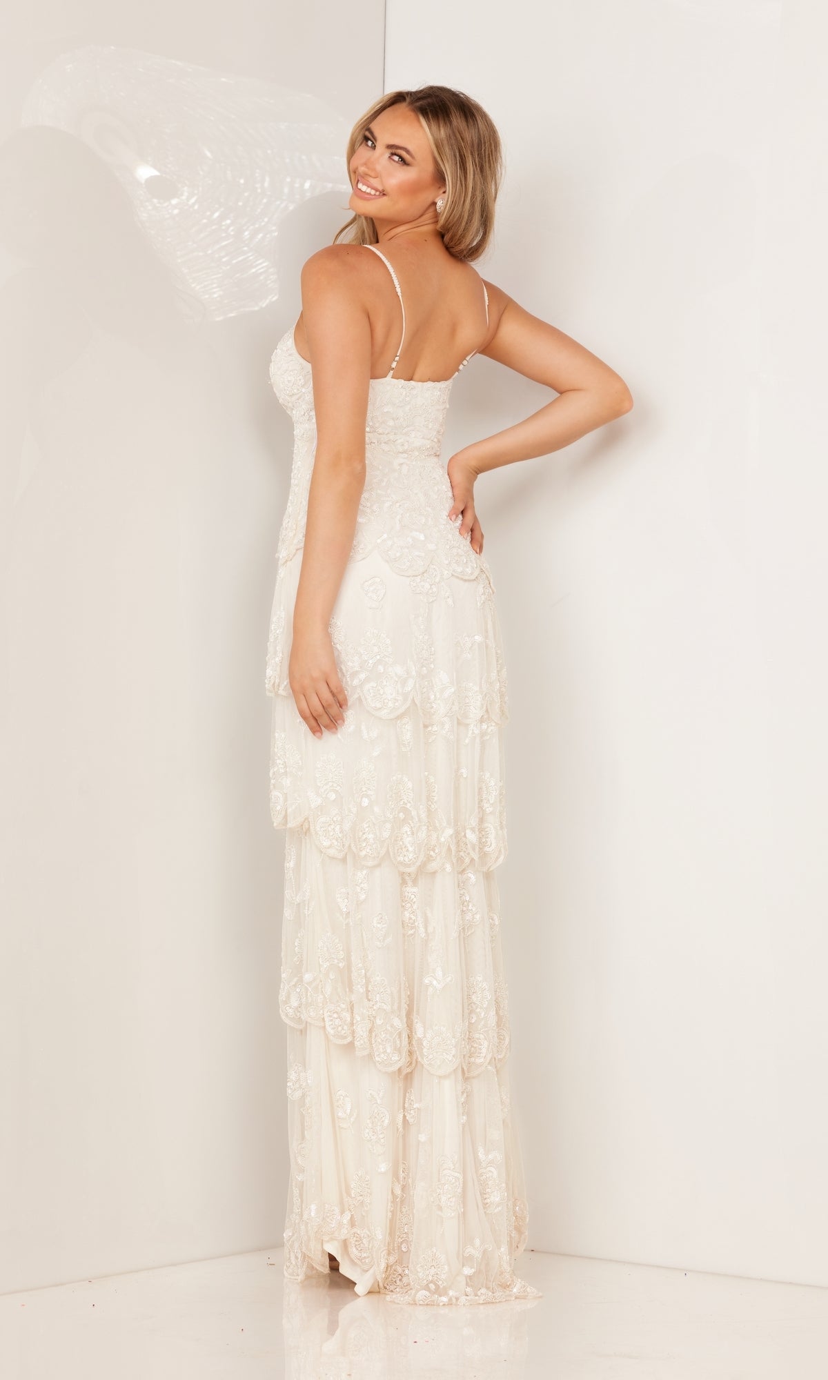 A woman wears the Long Beaded Prom Dress with Tiered Skirt 1190, a white sleeveless formal gown, facing away and smiling over her shoulder against a plain white background.