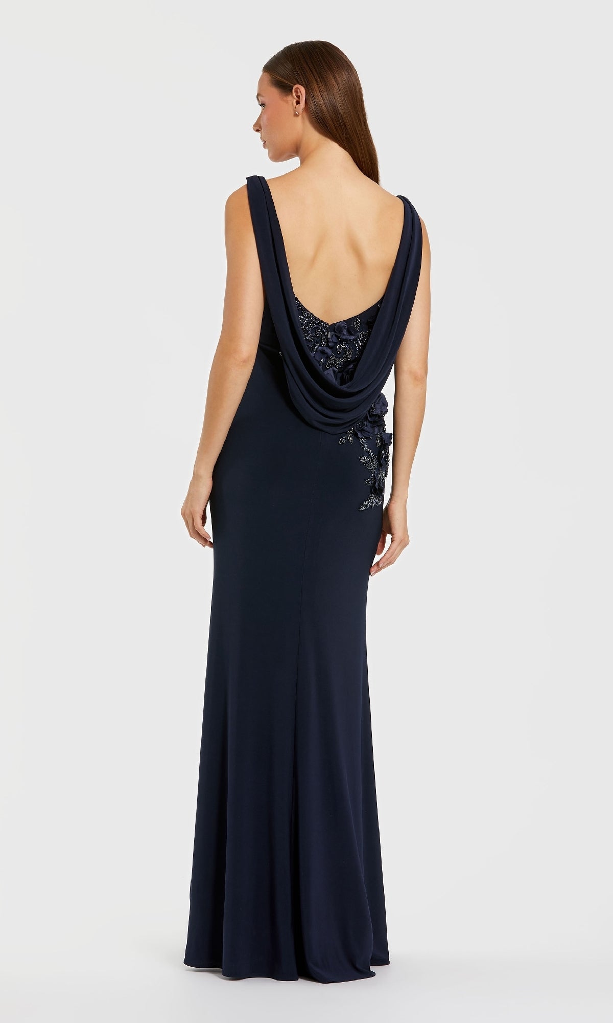 Woman in the Mac Duggal 11910 Cowl-Neck Long Formal Dress, featuring a navy blue hue, draped cowl back, and floral embellishments, standing against a plain light background.