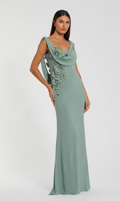 A woman models the Mac Duggal 11910 cowl-neck long formal dress in sage green, with floral embellishments on one side, posed against a plain white background.