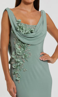 A woman wears the Mac Duggal 11910 Cowl-Neck Long Formal Dress, a sleeveless sage green gown featuring a draped neckline and floral appliqué on the bodice and side.