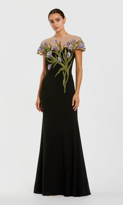 A woman wears the Mac Duggal 11912 Long Beaded-Tulip Formal Dress, a black gown with sheer short sleeves and floral embroidery in purple and green on the bodice and upper skirt.