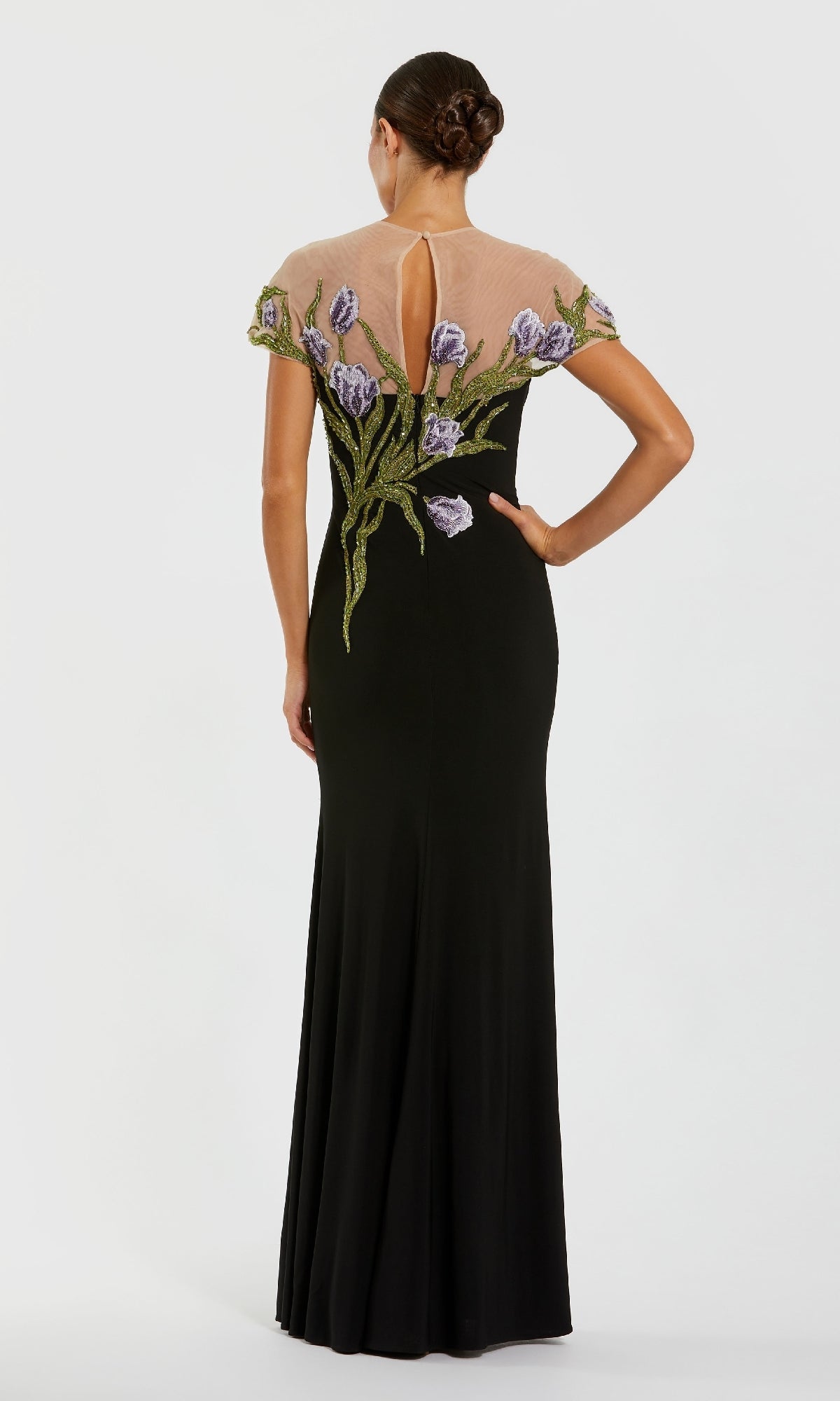 A woman in a Mac Duggal 11912 Long Beaded-Tulip Formal Dress stands facing away, her hair in a neat bun. The black gown features sheer shoulders and floral embroidery on the upper back.