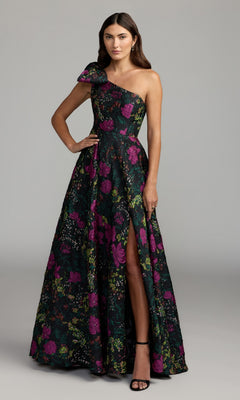 Wearing the One-Shoulder Floral Formal Gown Mac Duggal 11919 in Black Multi—a striking floor-length black dress adorned with pink and green floral patterns and featuring a thigh-high slit—a woman stands elegantly against a plain gray background.