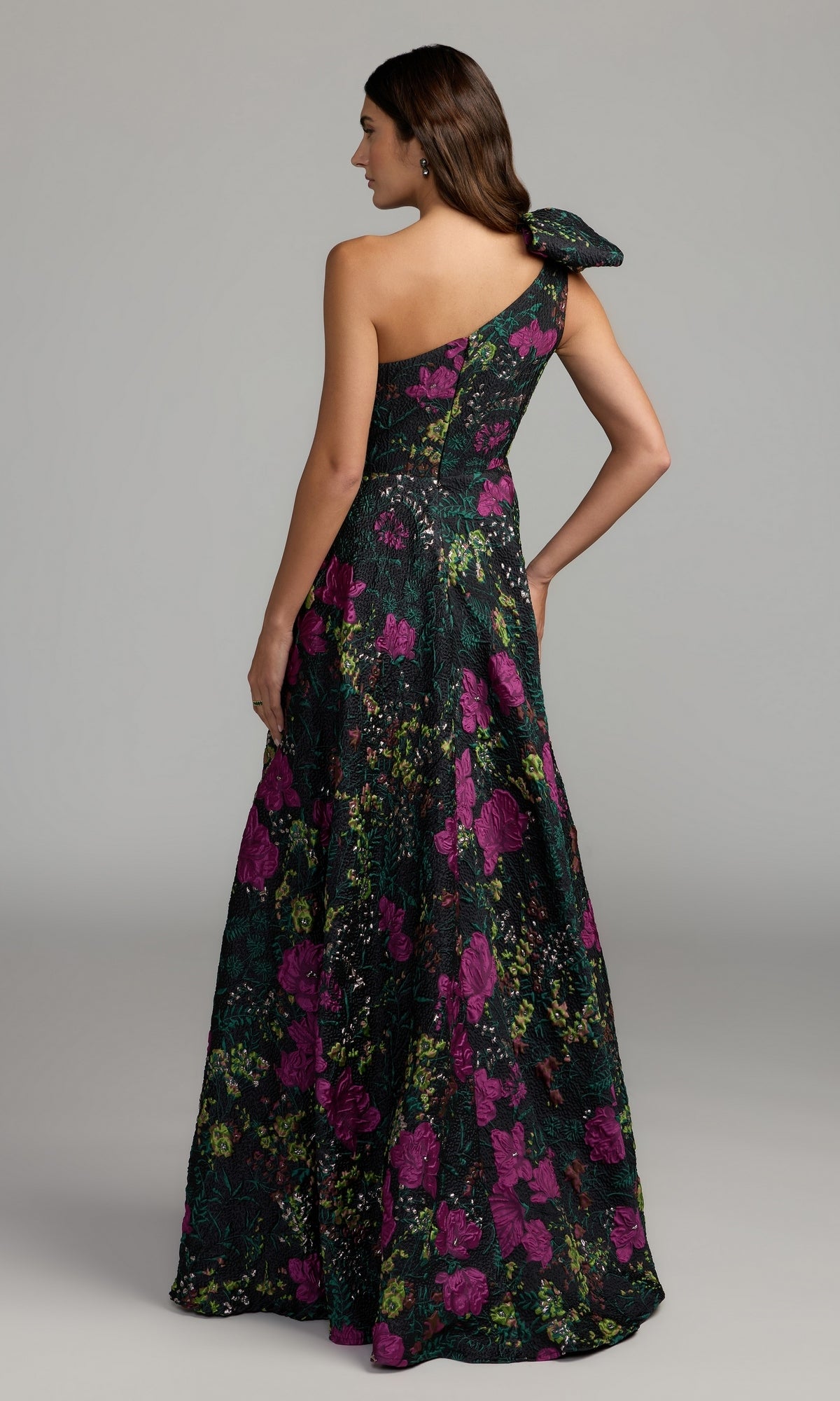 A woman stands facing away, wearing the Mac Duggal 11919 One-Shoulder Floral Formal Gown—a striking dress with a dark green base, bright pink and purple floral patterns, and a dramatic large bow—making it the perfect choice for any special occasion.