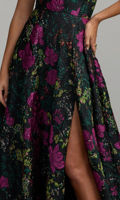 Wearing the One-Shoulder Floral Formal Gown Mac Duggal 11919—an elegant black dress detailed with purple floral patterns and a thigh-high slit—the person stands confidently with one hand on their hip, showcasing a stunning choice for prom or any formal occasion.