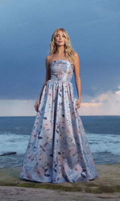 A woman in the Mac Duggal 11921 long formal dress, a strapless blue floral ballgown, stands on a rocky shore as its pastel hues echo the ocean and cloudy sky.
