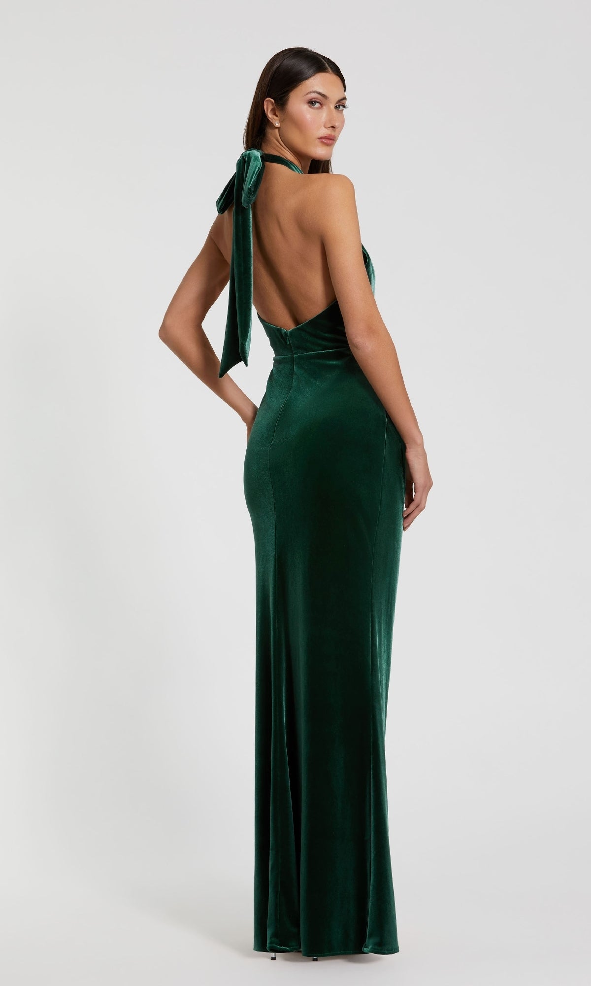 Woman wears the Green Velvet Halter Formal Dress: Mac Duggal 11924—an emerald green floor-length halter gown with open back and bow detail—standing against a plain white background.