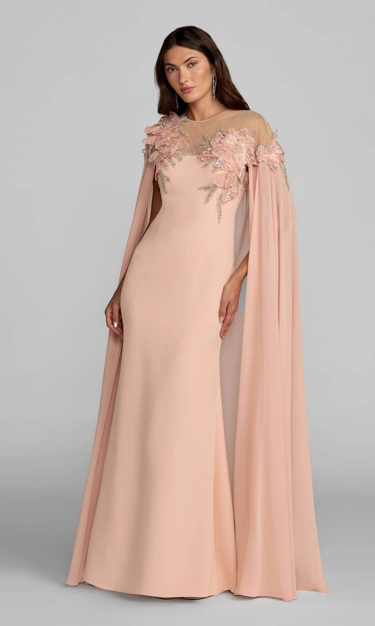 A woman stands against a plain gray background, wearing the Mac Duggal 11942 long formal dress in blush, featuring exquisite 3D floral appliques and attached cape sleeves.