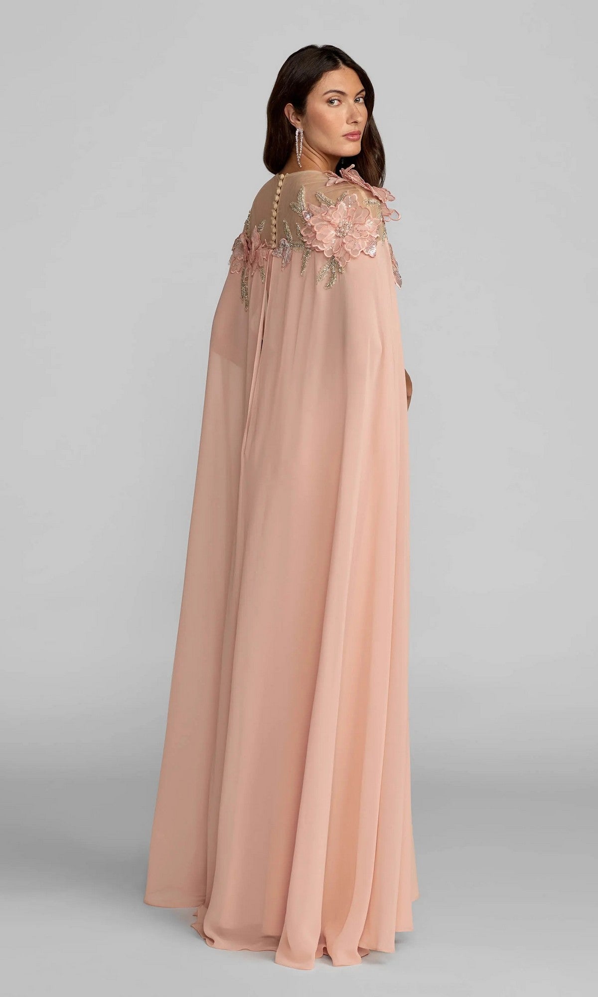 A woman wearing the Long Formal Dress: Mac Duggal 11942 in light pink, featuring a flowing silhouette with sheer detailing and intricate 3D floral appliques on the shoulders, stands against a plain gray background.