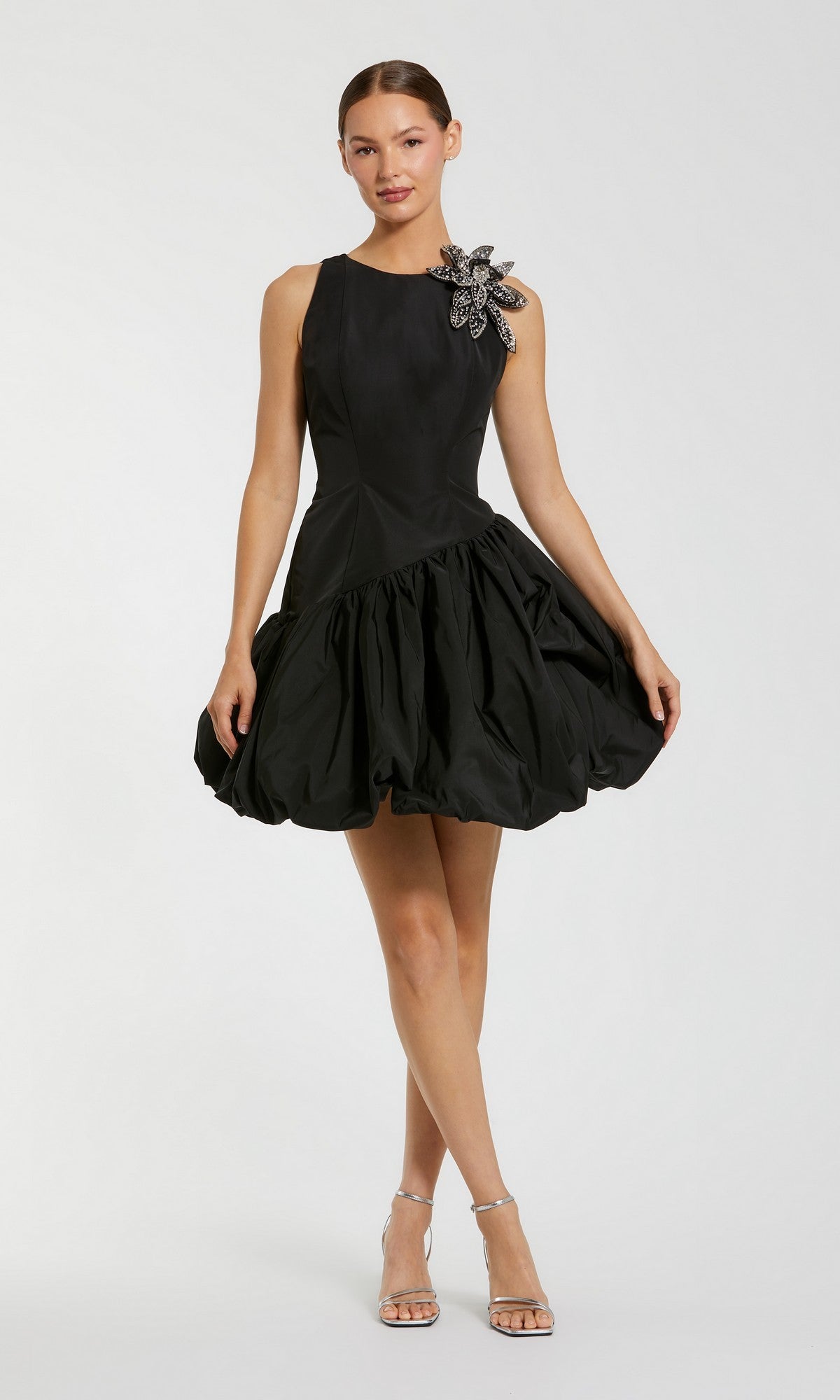 A woman stands against a plain light background wearing the Mac Duggal 11952 Short Cocktail Party Dress in black—a sleeveless mini dress with a flared bubble hemline and beaded flower detail on one shoulder—paired with silver high-heeled sandals.