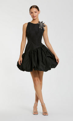 A woman stands against a plain light background wearing the Mac Duggal 11952 Short Cocktail Party Dress in black—a sleeveless mini dress with a flared bubble hemline and beaded flower detail on one shoulder—paired with silver high-heeled sandals.