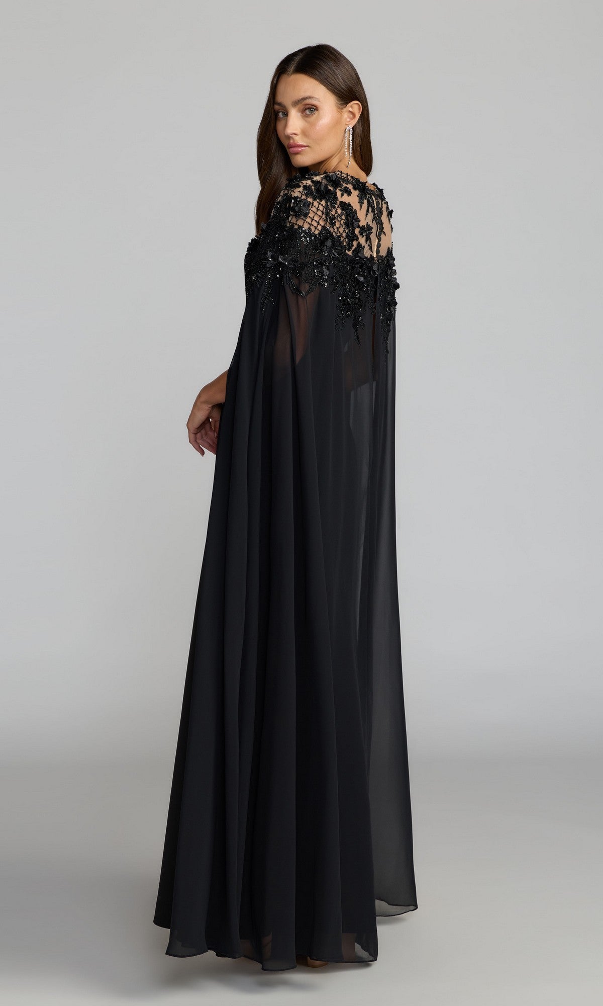 A woman models the Long Formal Dress: Mac Duggal 11954—a black crepe gown featuring chiffon cape sleeves and sequin floral embroidery on the upper bodice—as she stands against a plain studio background.