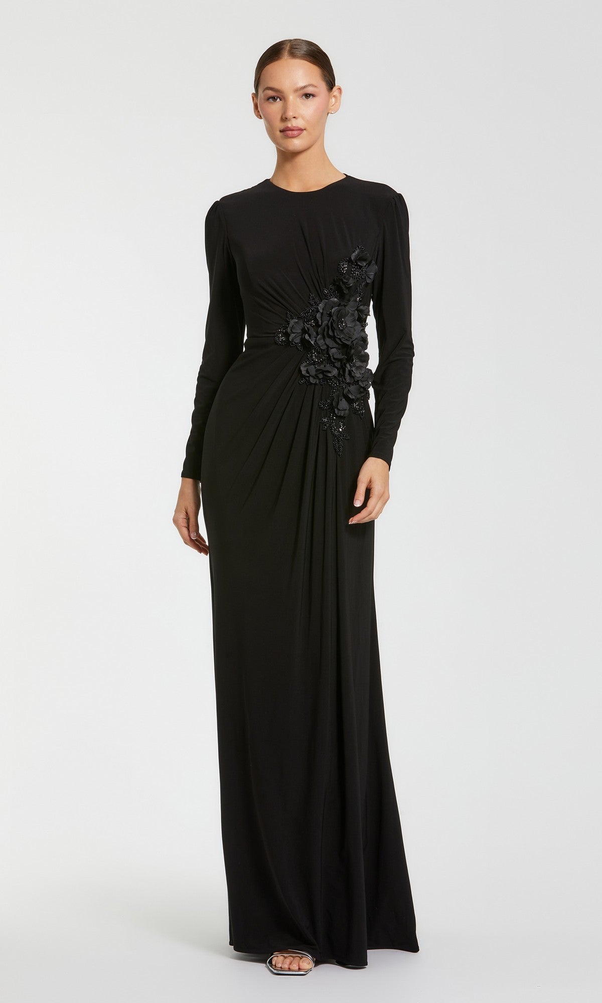 A woman stands wearing the Mac Duggal 11960 long formal dress in black—a modest gown with long sleeves, floral embellishments at the waist, a floor-length skirt, and open-toe heels.