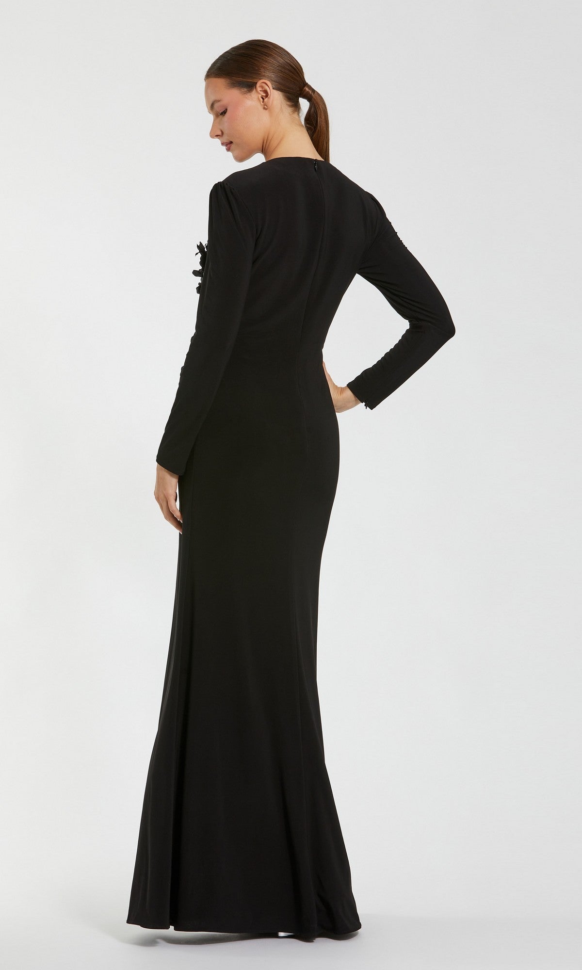 A woman with light skin and her hair tied back in a low ponytail stands with her back to the camera, wearing the Mac Duggal 11960 Long Formal Dress. The dress is black, long-sleeved, and floor-length. The background is plain white.