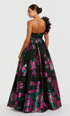 Woman wearing the Mac Duggal 11967 long formal dress—a black floral ballgown with pink and green flowers and a ruffled one-shoulder strap—posing against a plain light background, exuding classic elegance.