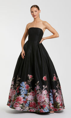 A woman stands against a plain background wearing the Mac Duggal 11970 Strapless Floral Prom Ball Gown, a long black dress with a full skirt adorned with pink and blue floral patterns near the hem.