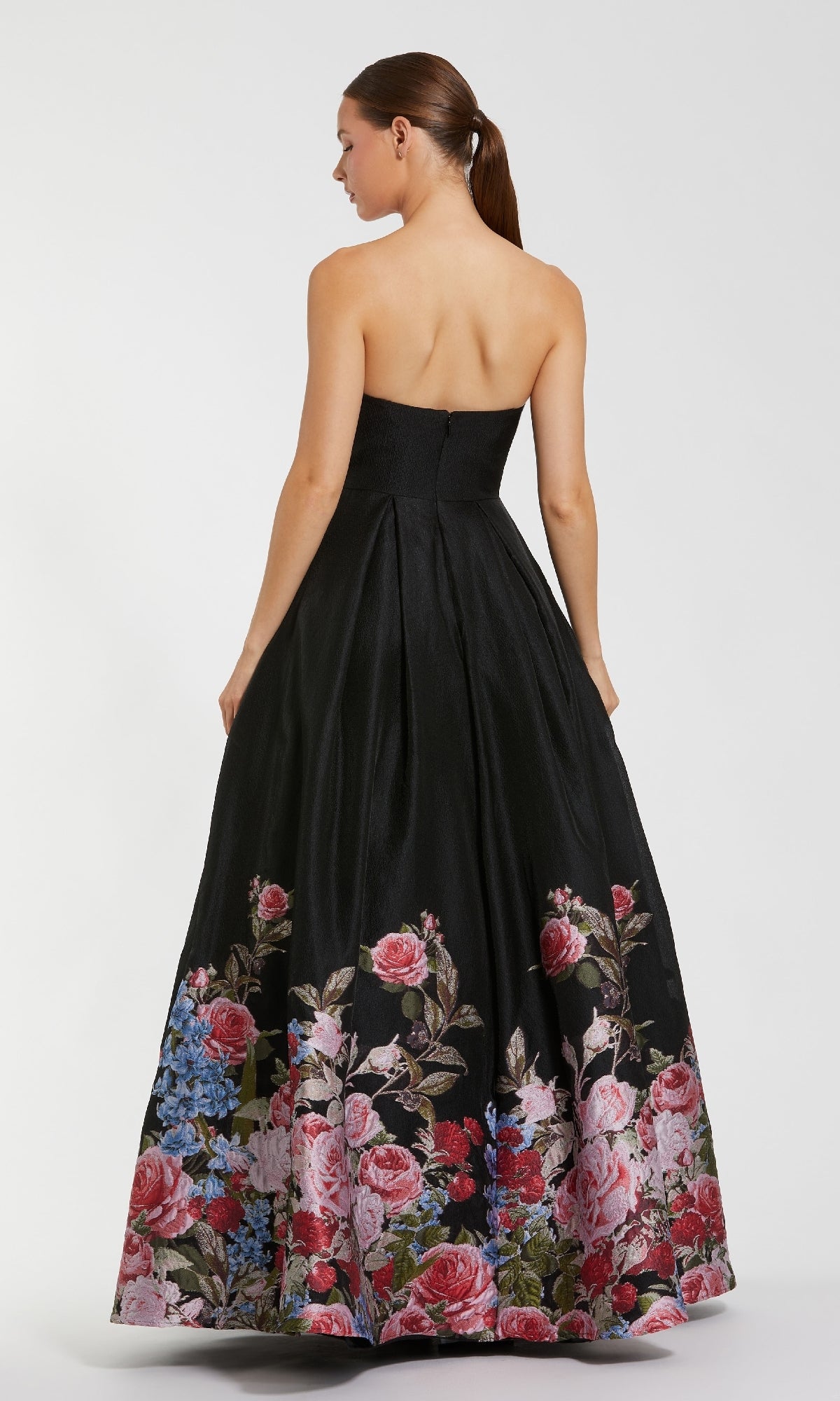 A woman models the Mac Duggal 11970 Strapless Floral Prom Ball Gown, a long black A-line dress with pink, red, and blue floral accents near the hem. The elegant back view highlights its strapless design and flowing skirt.