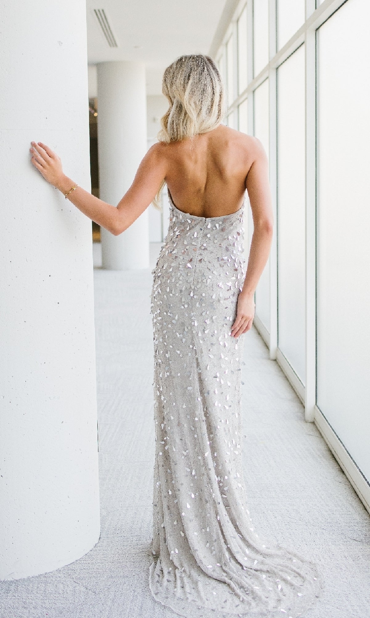 A woman wearing the Aleta Halter Beaded Champagne Prom Dress 1197 stands facing away, resting one hand on a white column in a bright hallway with large windows.