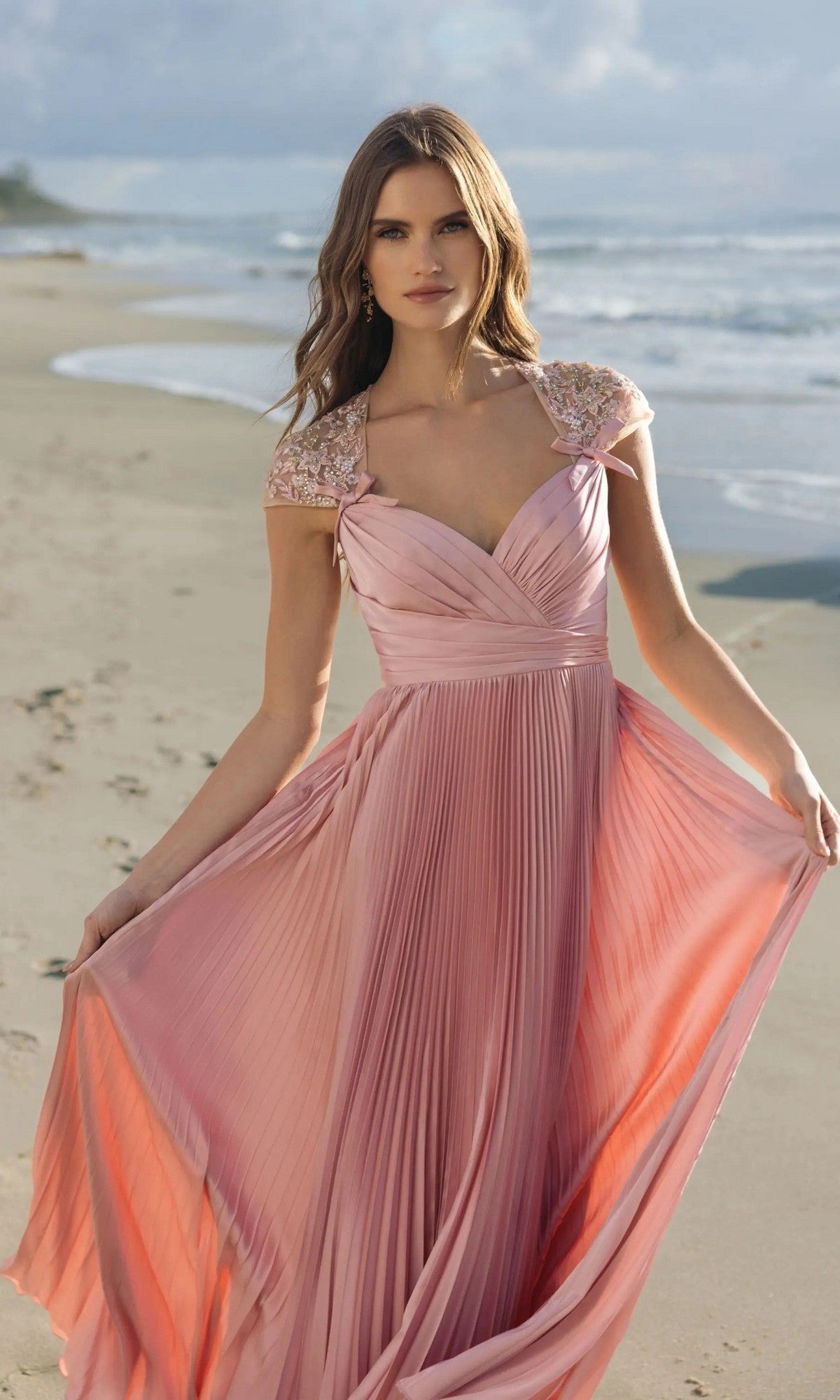 A woman wearing the Long Formal Dress: Mac Duggal 11987 in rose, featuring flowing pleats and embellished cap sleeves, stands on a sandy beach by the ocean.