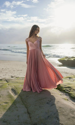A woman stands on a rocky beach in the Mac Duggal 11987 long formal dress, its pleated pink fabric glowing as waves crash and sunlight illuminates the scene.