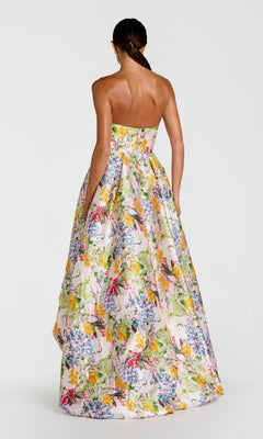 A woman faces away, wearing the Mac Duggal 11991 long formal dress—strapless with a vibrant floral print on a light background and featuring a dramatic high-low bubble hem.