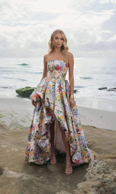 A woman stands on a rocky beach in Mac Duggal 11991, a strapless floral Mikado long formal dress with ruffled layers and a high-low bubble hem, set against a cloudy sky and ocean backdrop.