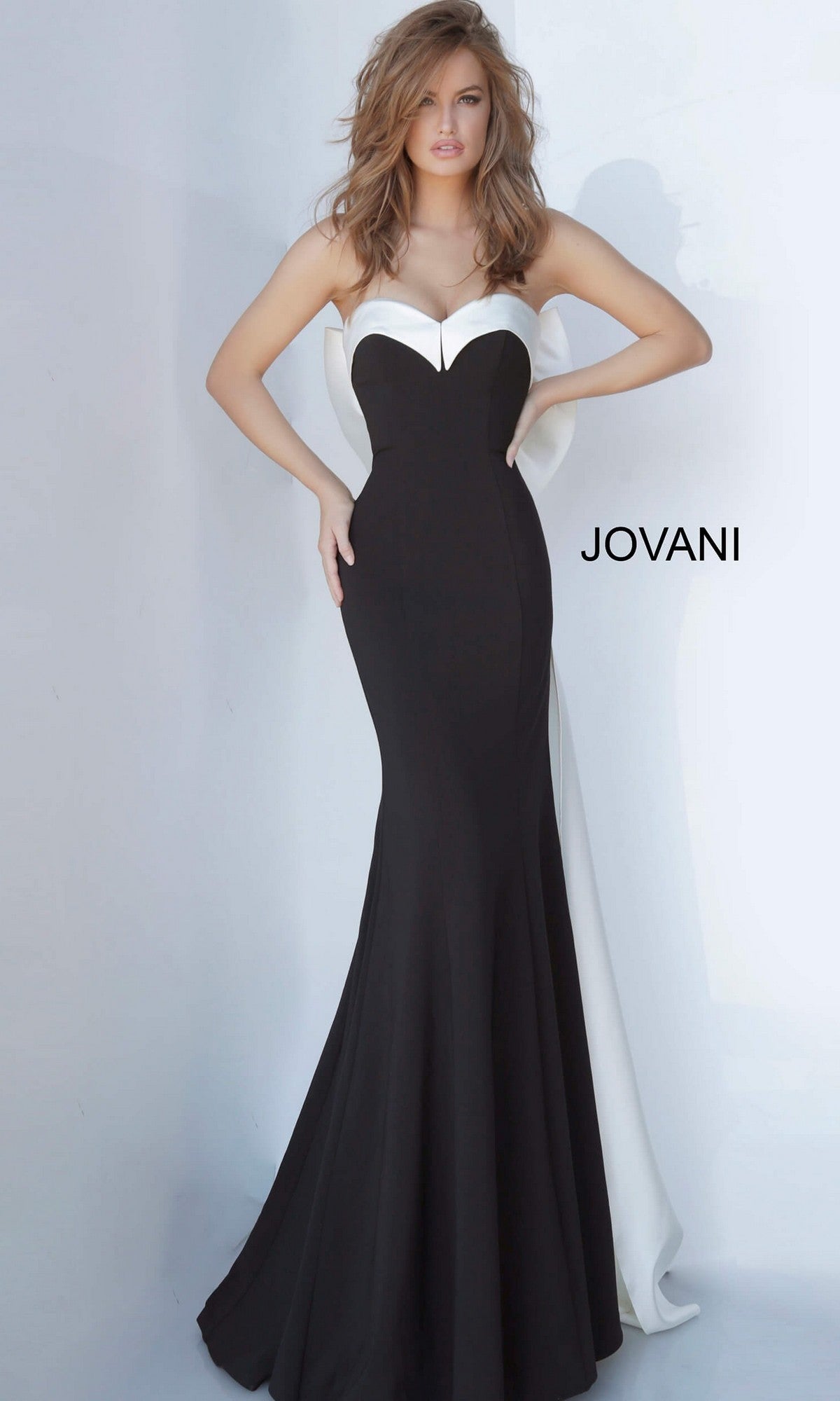 A woman stands against a plain light background wearing the Jovani 12020 long formal dress—a striking strapless satin gown in a black and white mermaid silhouette—with “JOVANI” text on the right.