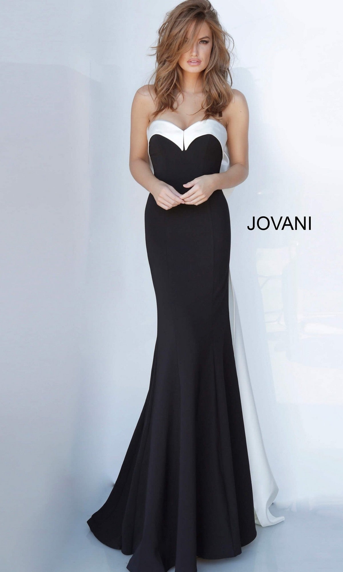 A woman stands against a light background, wearing the Jovani 12020 long formal dress—a black and white strapless satin gown with an elegant mermaid silhouette. The word “JOVANI” appears to the right of her.