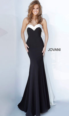 A woman stands smiling against a plain light background, wearing the Jovani 12020 long formal dress. This strapless, floor-length gown features a striking black and white satin design with an elegant mermaid silhouette and a dramatic oversized bow at the back. The word “JOVANI” appears to the right.