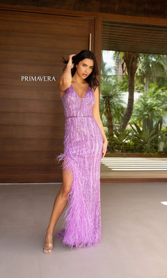 A woman in the Primavera Feathered Long Sequin Prom Dress 12108, a sleeveless lilac gown with beadwork, feather accents, and a thigh-high slit, stands before a wooden door labeled "PRIMAVERA.