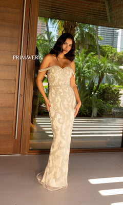 A woman stands before glass doors in the Primavera Beaded Off-the-Shoulder Prom Dress 12110, a long cream gown adorned with intricate silver embellishments. The word "PRIMAVERA" appears on the left.