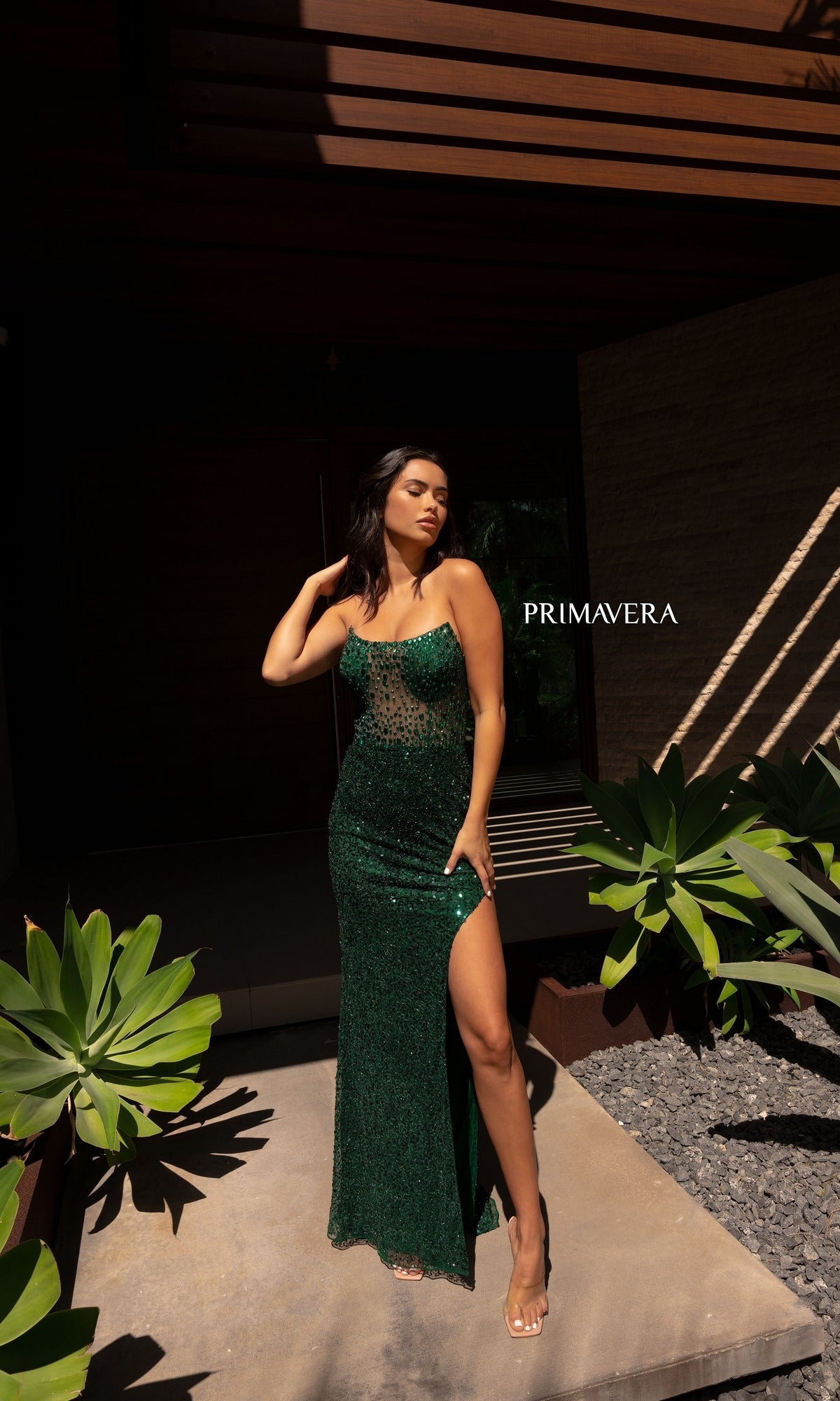A woman in the Primavera 12123 Strapless Long Beaded Prom Dress with a thigh-high slit stands outdoors by plants, casting a shadow. 