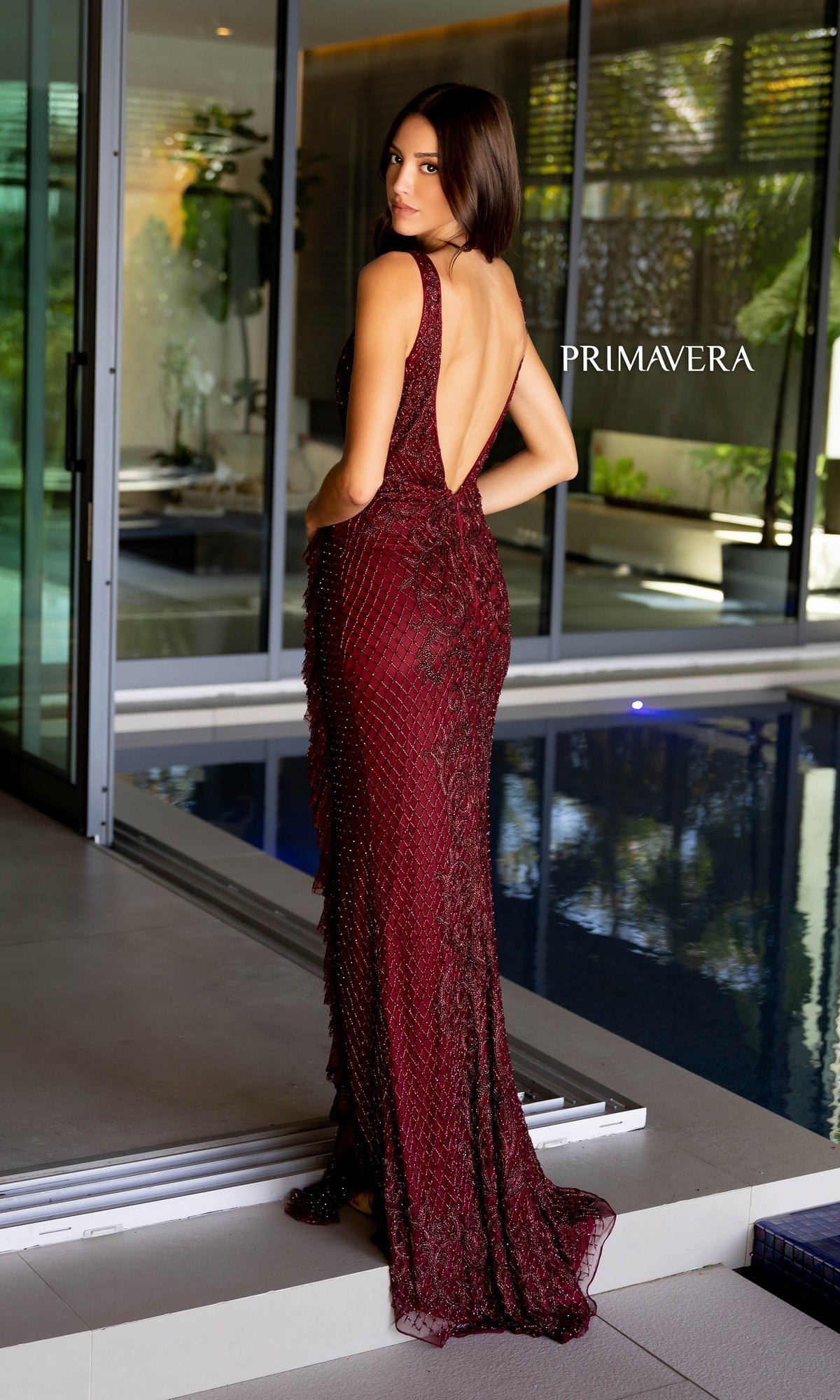 A woman in the Long Prom Dress 12160 by Primavera, a sleeveless deep red beaded formal gown with a v-neckline and backless design, stands poolside looking over her shoulder. The word 