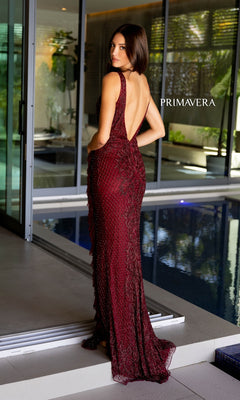 A woman in the Long Prom Dress 12160 by Primavera, a sleeveless deep red beaded formal gown with a v-neckline and backless design, stands poolside looking over her shoulder. The word "Primavera" is visible on the dress.