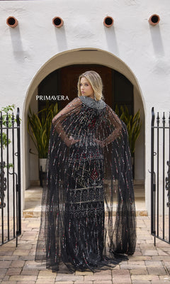 A woman wears the Primavera 12172 long formal dress with a sheer, beaded cape, standing with her back to an arched doorway framed by black gates and potted plants.