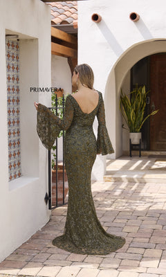 A woman wearing the Primavera 12185 long formal dress in olive green, featuring beads, bell sleeves, and flared cuffs, stands in a sunlit courtyard facing away from the camera.
