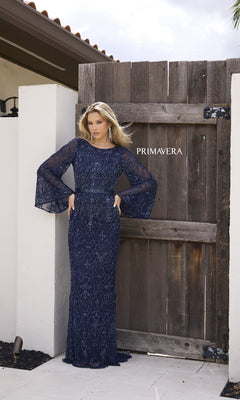 A woman in the Primavera 12187 long formal dress—a navy blue lace gown with beaded accents and long bell sleeves—stands before a wooden gate labeled "PRIMAVERA.