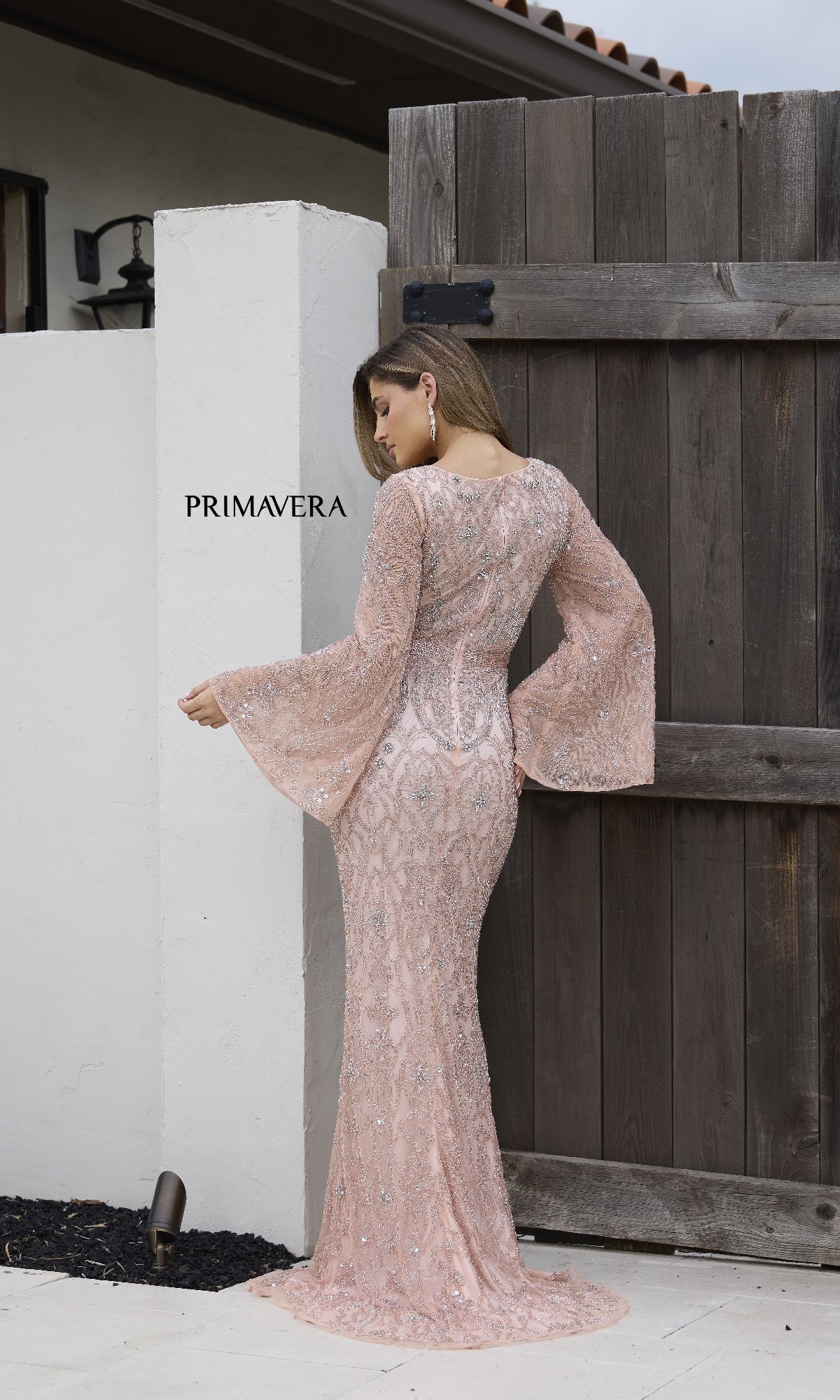 A woman wearing the Primavera 12187 long formal dress with long bell sleeves and light pink sequins stands outdoors in front of a wooden gate, facing away from the camera.